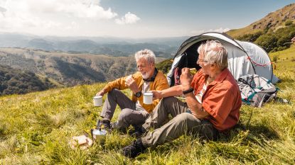 Two older men laugh and drink coffee while sitting outside their tent in the mountains.