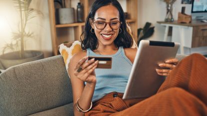 A woman sitting on her sofa smiles as she looks at her credit card and holds a tablet.