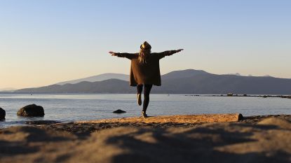 A woman balances on one foot while facing a lake and mountains.